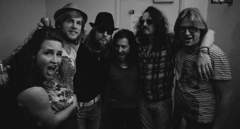Milkbar backstage, zes bandleden lachend met de armen om elkaar heen
