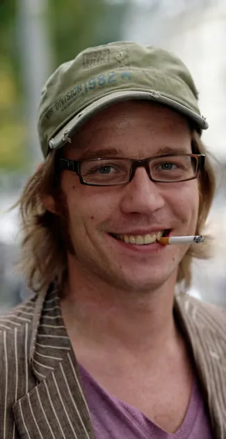 Bandlid van Milkbar met pet, bril en sigaret, lachend in de camera